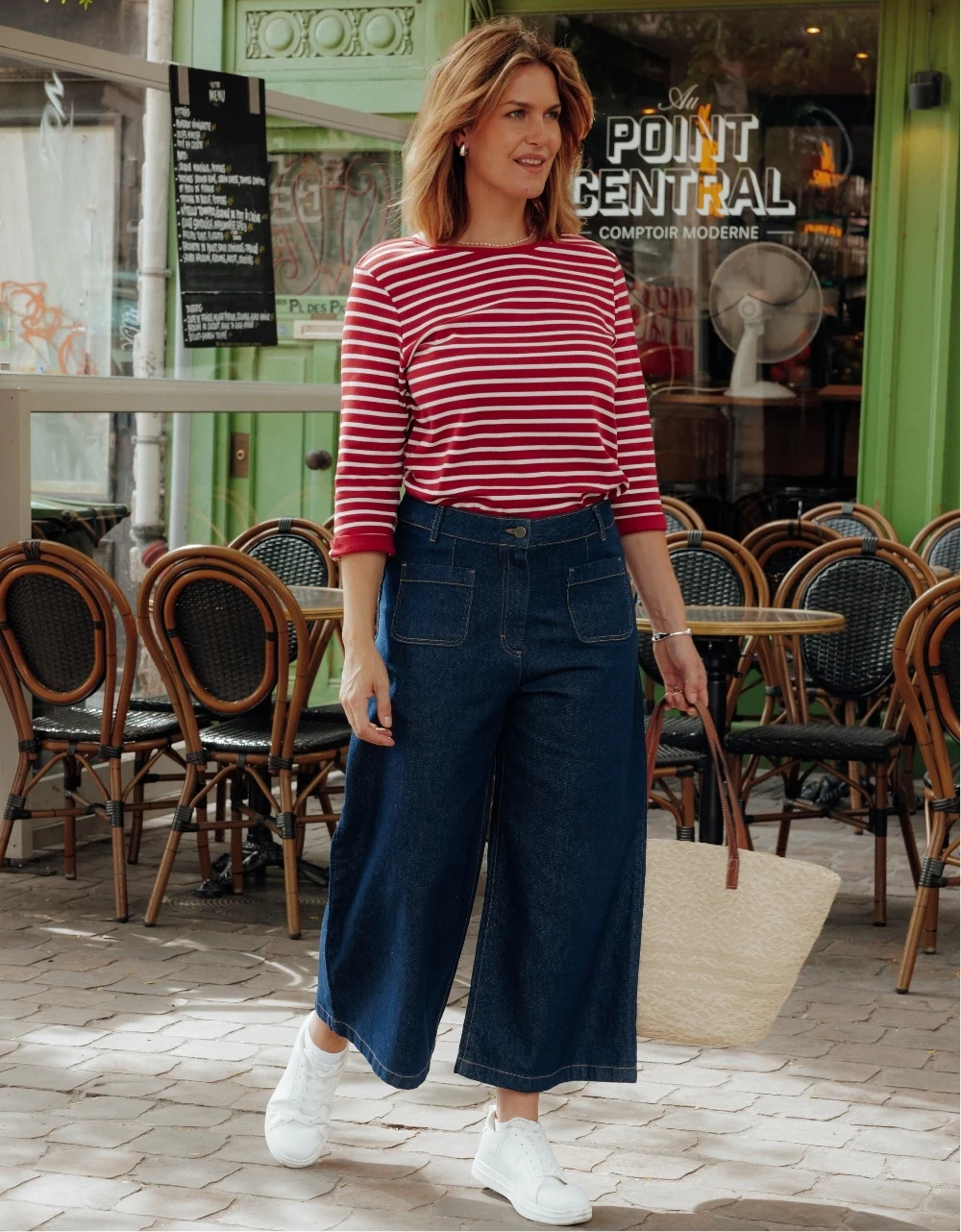 Mannequin en marche devant un café, portant une marinière rouge, un jean large bleu et des baskets blanches Christine Laure.