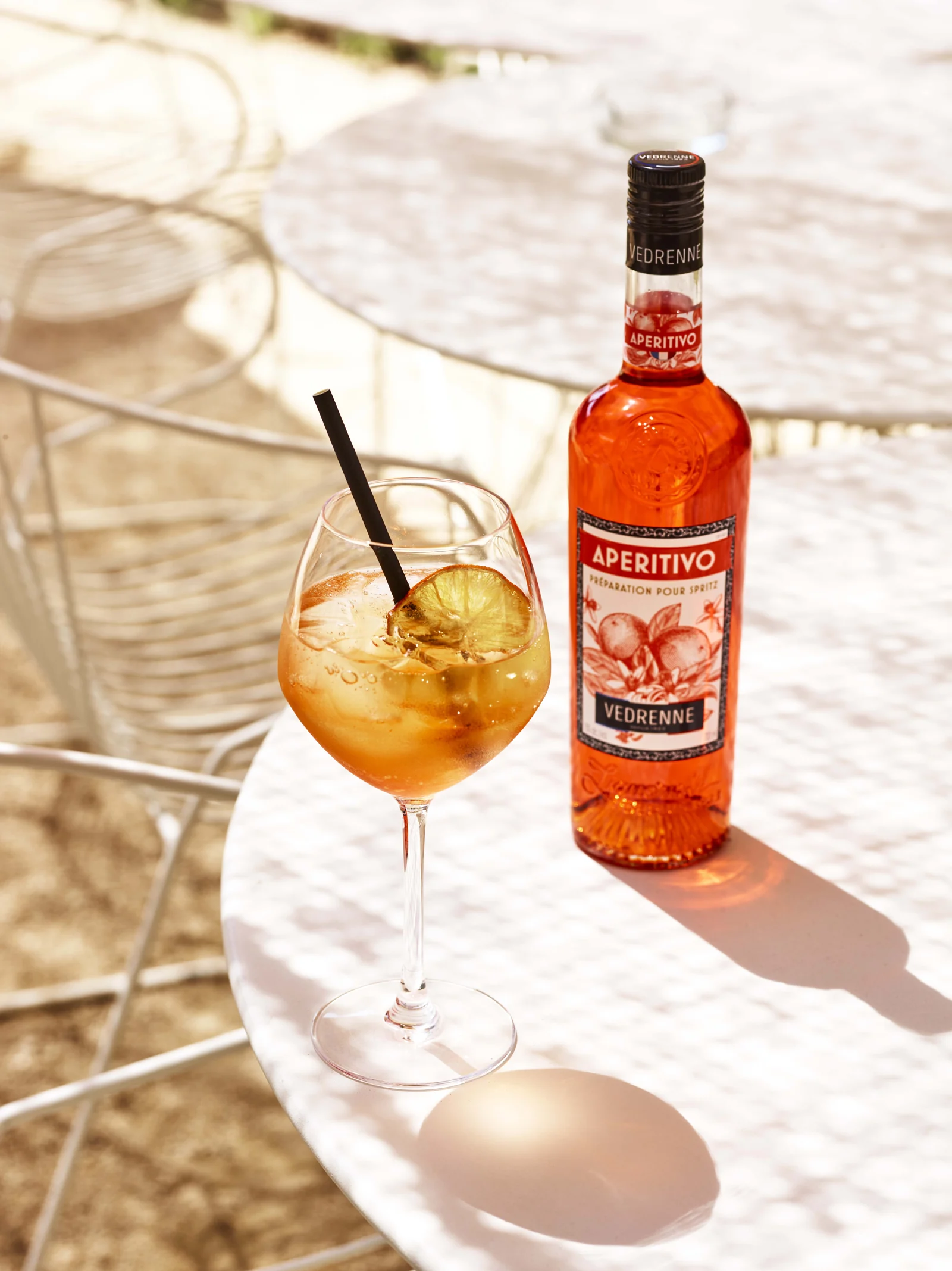 Bouteille Aperitivo Vedrenne et cocktail orangé sur une table de terrasse ensoleillée.