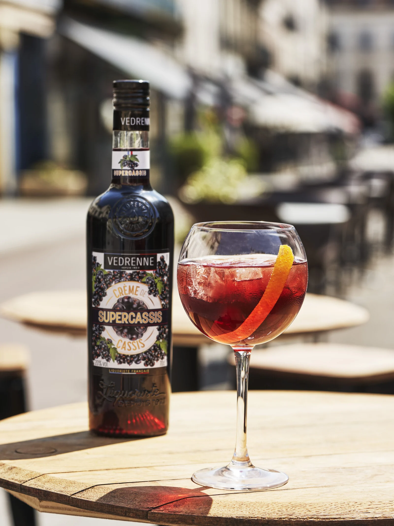 Bouteille de Supercassis Vedrenne et cocktail rouge sur une table de terrasse.